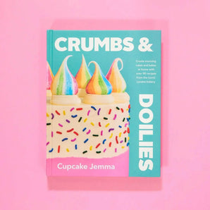 The Crumbs & Doilies Recipe Book with colorful cake on cover by Cupcake Jemma on pink background.